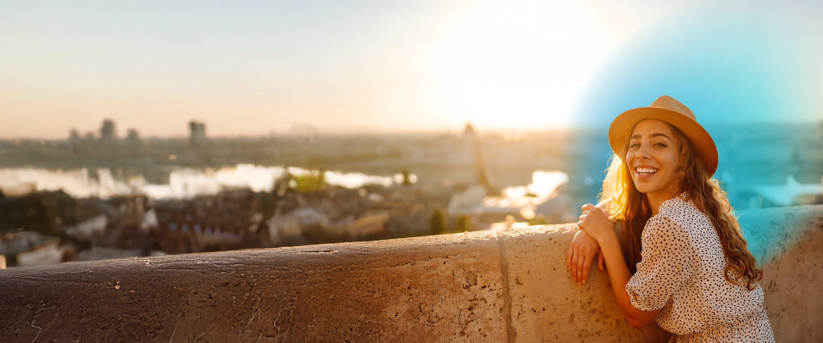 Young woman wearing a white polka dot dress and tan hat standing on a bridge admiring a foreign city in the background 