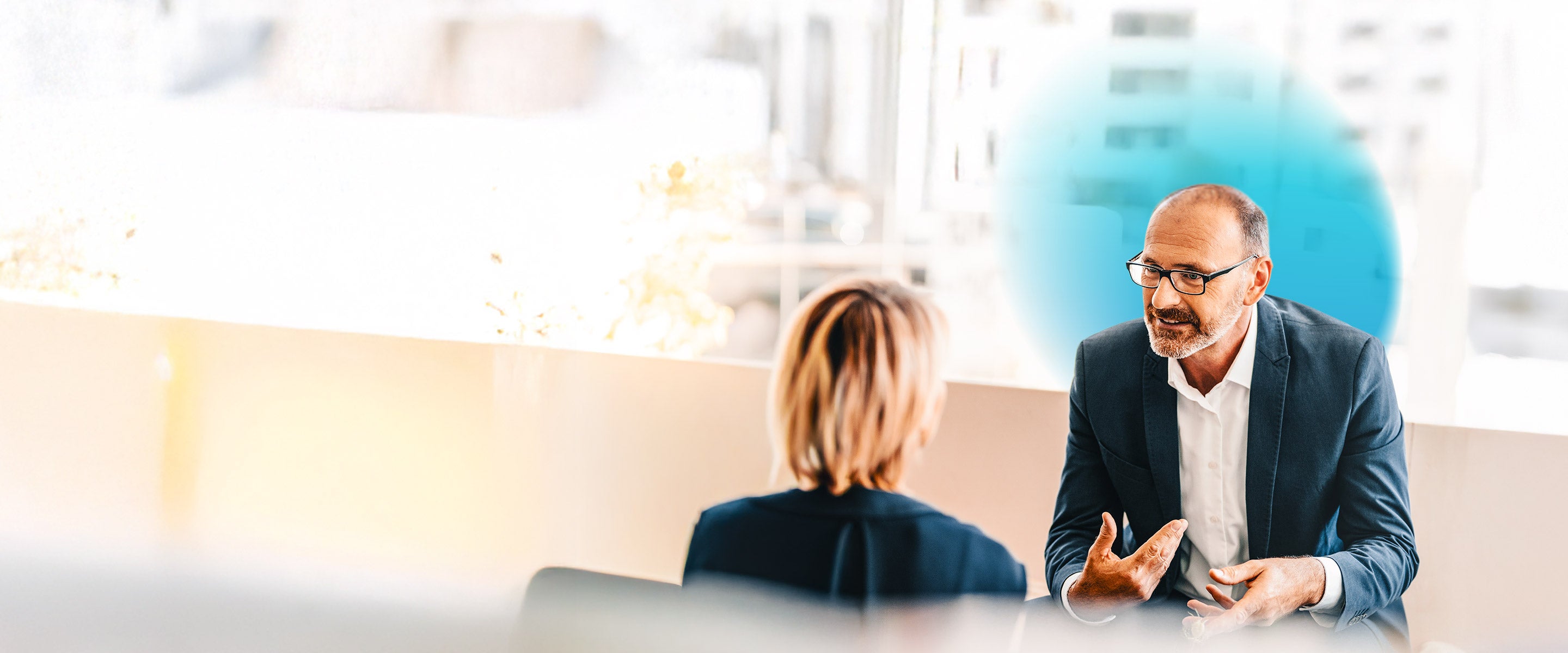 Businessman wearing glasses sitting down and the background is blurred with a blue gradient hovering around him while talking to a woman facing him