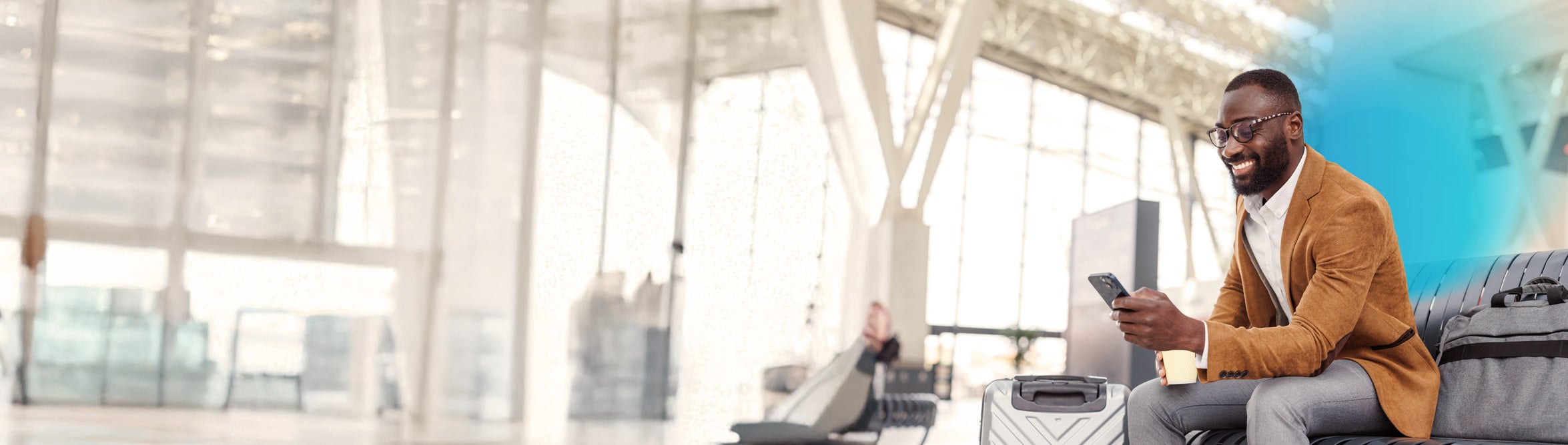 Smiling man wearing glasses and a brown blazer sitting at the airport with a suitcase and looking at his phone with a blue gradient over him