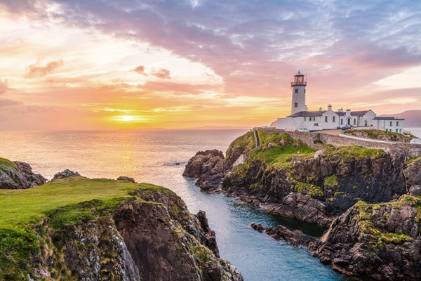 Cliffs in Ireland covered by green grass with a blue sea in the middle and a white lighthouse on the right-hand cliff as the sun sets in the sky