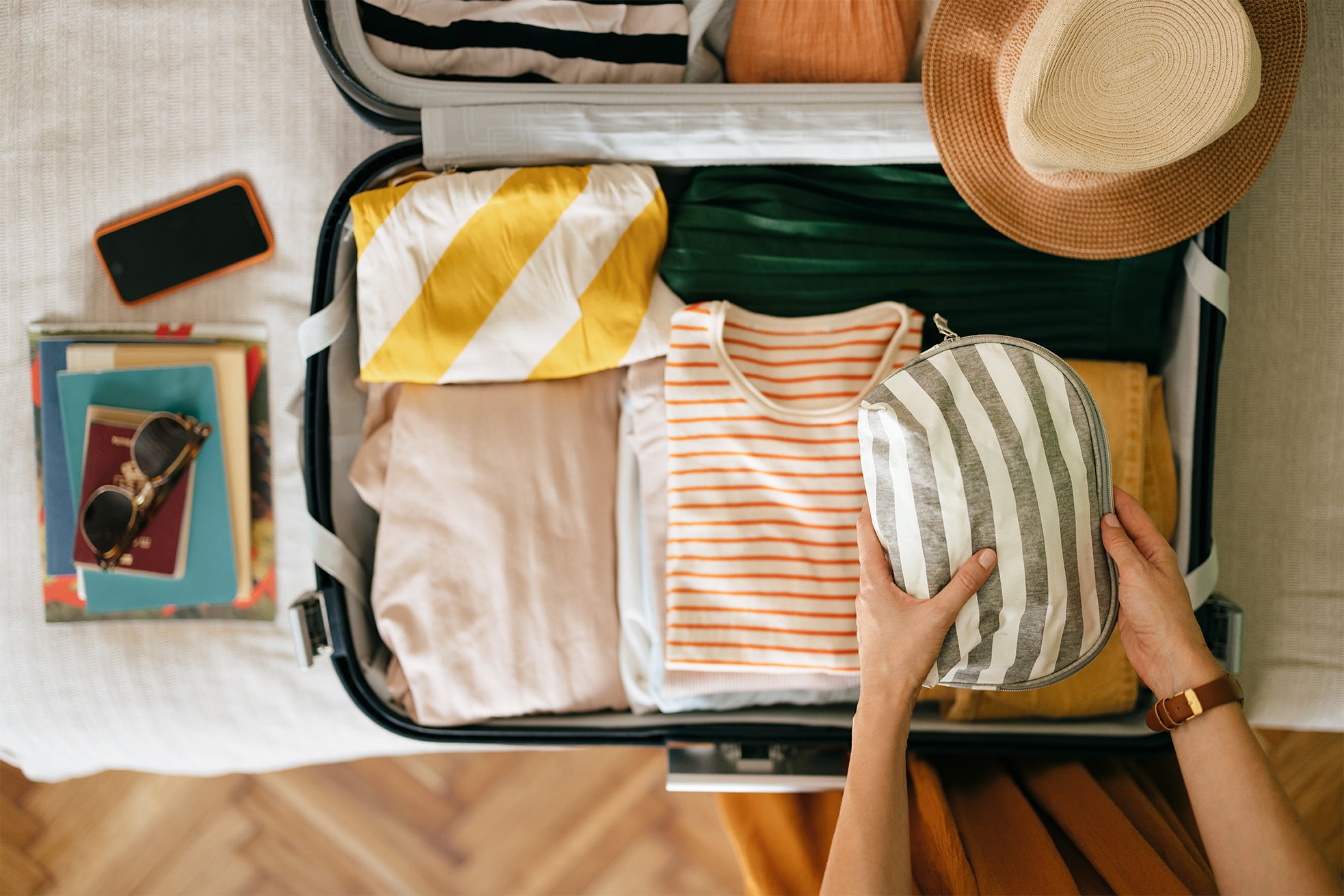 A woman holding a grey and white stripped makeup bag over top of an open suitcase filled with orange, yellow and green clothes with books and her phone to the left on her bed 