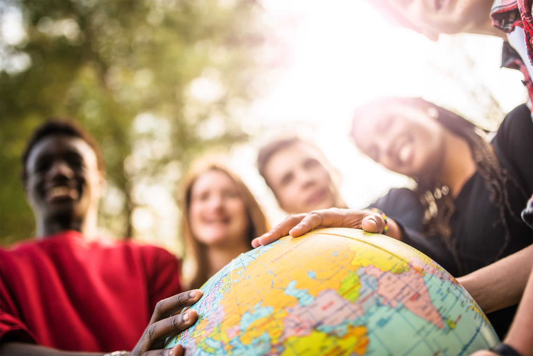 Group of young multiethnic college students holding a globe of the world with trees in the background and sunlight coming through 