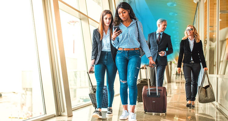 Two female friends and a businessman and woman walking behind them all rolling suitcases in an airport by a big window and blue gradient hovering around them 