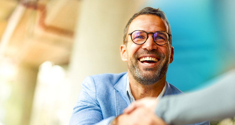 Smiling Caucasian man wearing a light blue blazer and glasses shaking someone's hand with a blurred background 