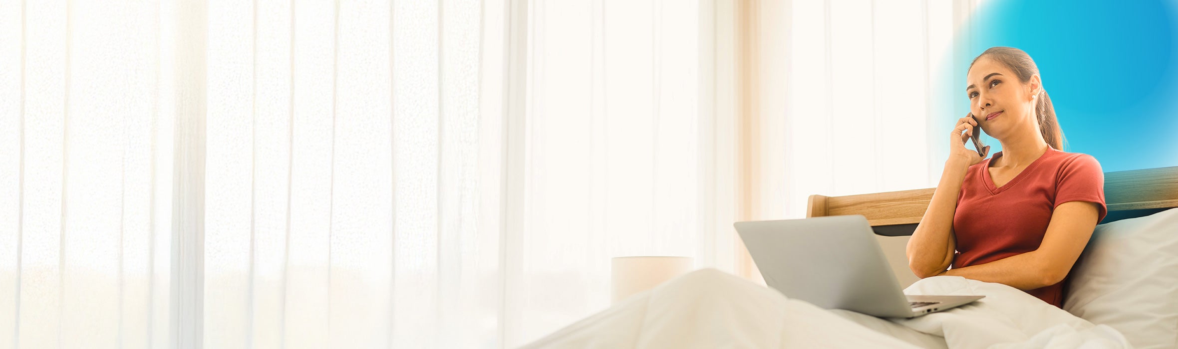 Woman wearing a rusty red t-shirt with her hair in a ponytail sitting in bed with her laptop and talking on her cell phone 