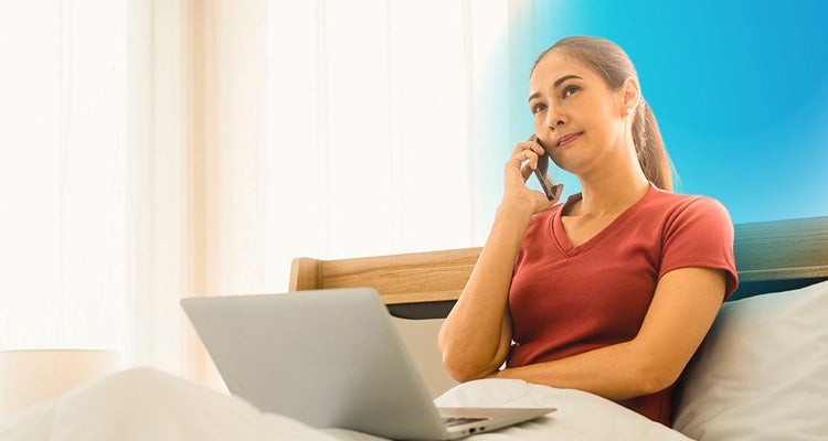 Woman wearing a rusty red t-shirt with her hair in a ponytail sitting in bed with her laptop and talking on her cell phone 
