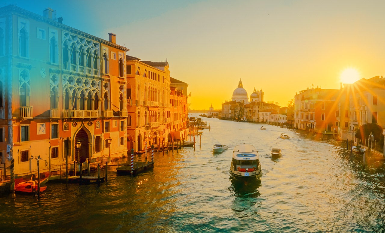Boat traveling the glittering waters of the Venice Canals surrounded by historic buildings lit up by the golden afternoon sky. 
