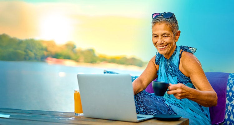 Senior Hispanic woman with short hair wearing a turquoise sarong drinking coffee and working on her laptop on her patio overlooking a tropical ocean 