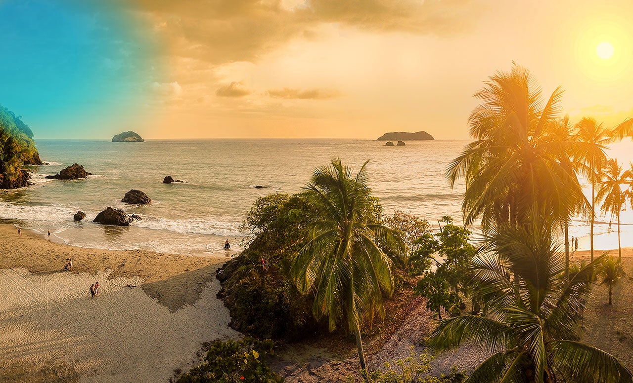 Tropical beach with palm trees, sand and mountains covered in green plants with large boulders in the ocean lit up by a golden sky 