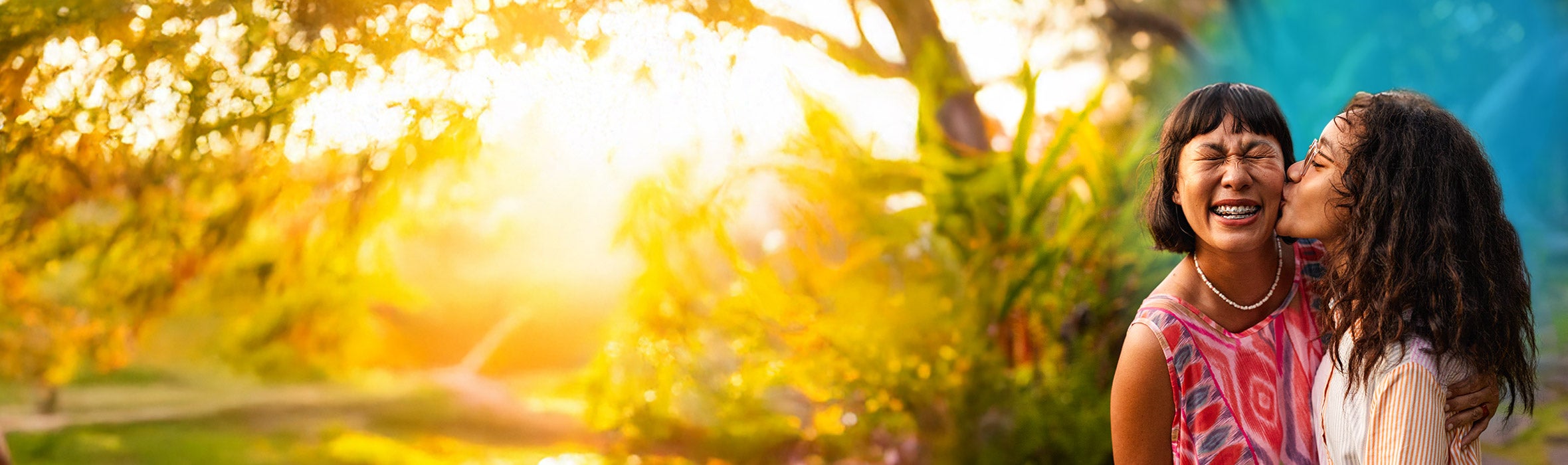 Young daughter kissing her mother on the cheek with a blue gradient hovering around them in a garden with the sun shining bright behind them 