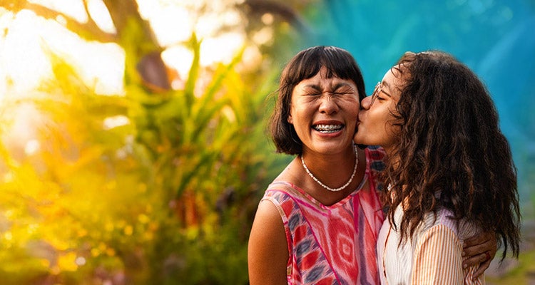 Young daughter kissing her mother on the cheek with a blue gradient hovering around them in a garden with the sun shining bright behind them 