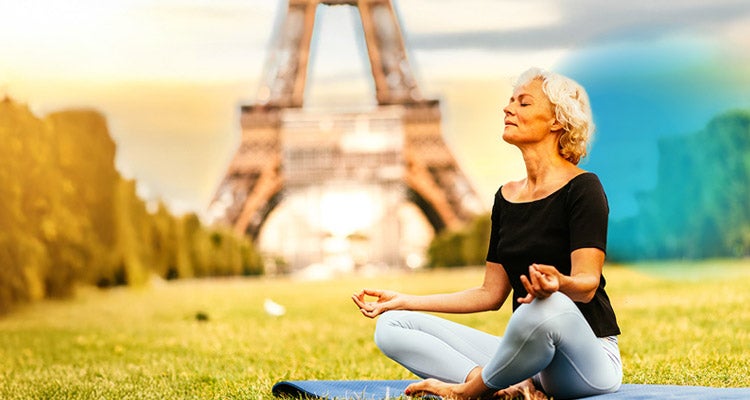 Older woman meditating in the grass on a yoga mat with a bridge and the sun shining in the background and a blue gradient hovering around her