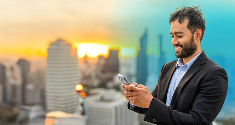 A businessman smiling while texting on his phone standing somewhere that is overlooking a modern city skyline with blue gradient hovering over man