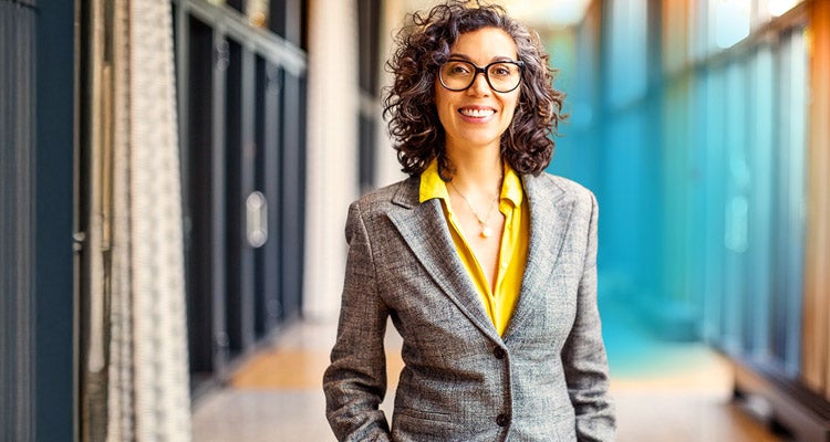 Middle aged Caucasian female professor with grey curly hair wearing a yellow blouse and grey blazer and glasses smiling in a school hallway 