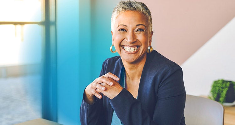 An African American businesswoman wearing a sharp navy suit with short silver hair and gold earrings smiling and sitting at her desk 