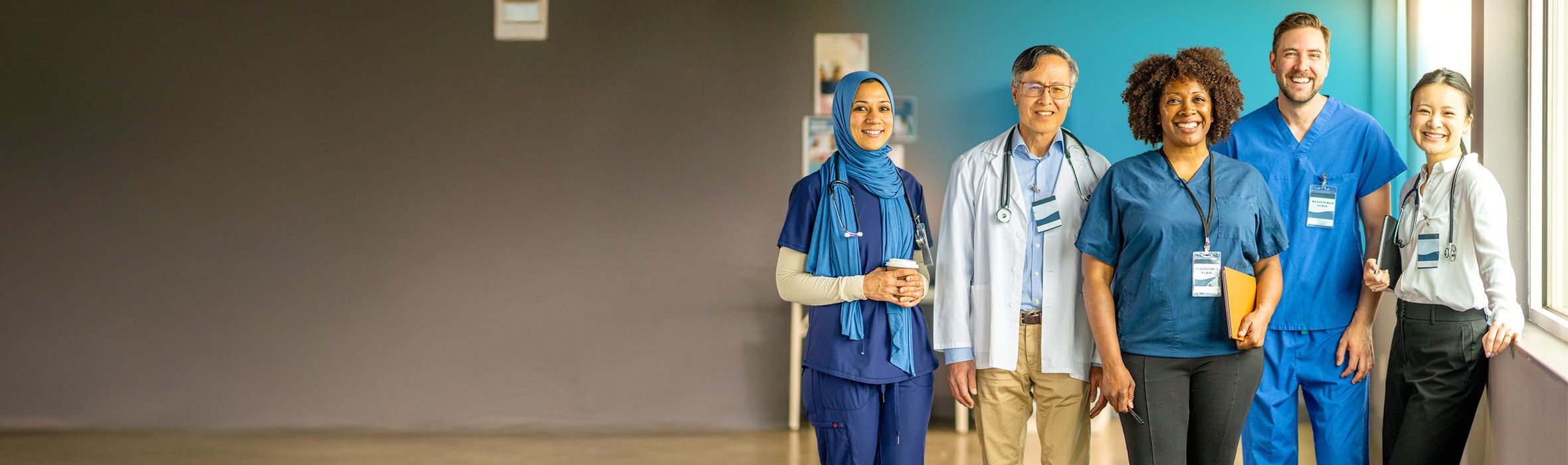 Multiethnic group of 5 medical professionals wearing blue, black and white scrubs and business attire smiling in a hospital corridor 