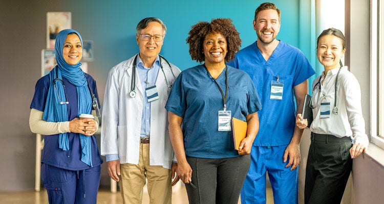 Multiethnic group of 5 medical professionals wearing blue, black and white scrubs and business attire smiling in a hospital corridor 