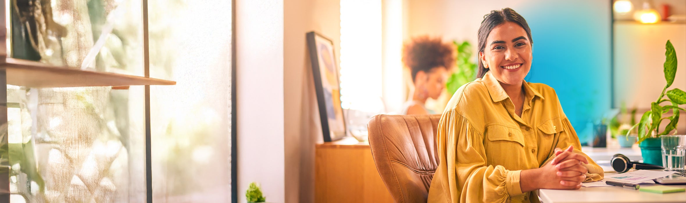 Young Hispanic businesswoman smiling wearing a yellow blouse sitting in a brown chair at her desk with sunlight coming through the window 