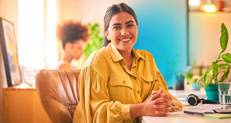 Young Hispanic businesswoman smiling wearing a yellow blouse sitting in a brown chair at her desk with sunlight coming through the window 