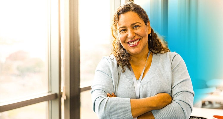 Hispanic woman teacher smiling wearing a grey sweater with her arms crossed standing in front of her desk in the classroom 