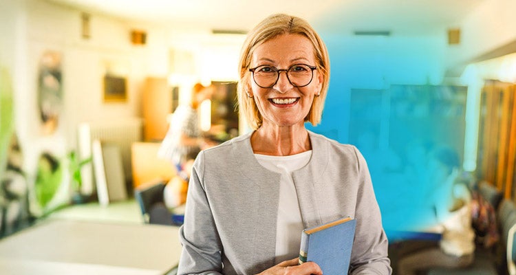 Senior Caucasian professor with short blonde hair and glasses and a grey cardigan standing in a classroom filed with plants, tables and sunlight  
