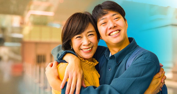 Middle-aged Asian mom wearing a yellow blouse hugging her college-aged son wearing a dark blue button up shirt inside of an airport 