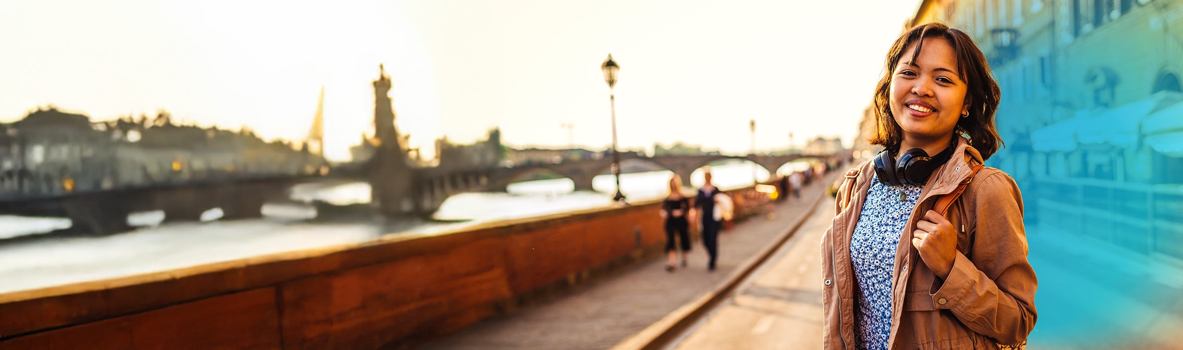 Young Asian woman wearing a brown jacket with headphones around her neck walking a scenic bridge in a foreign city 