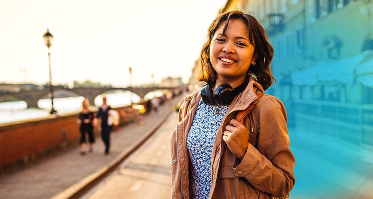 Young Asian woman wearing a brown jacket with headphones around her neck walking a scenic bridge in a foreign city 