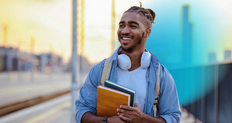 Young African American man smiling and wearing a light blue shirt and jacket holding college books walking in a college town. 
