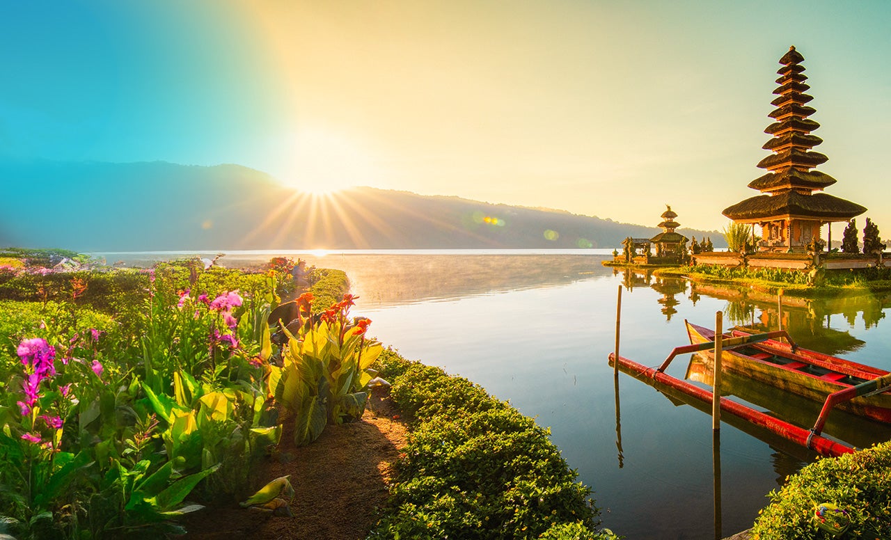Asian Temple on serene water lit up by a golden sunrise coming up from the mountains surrounded by greenery and bright purple and orange flowers 