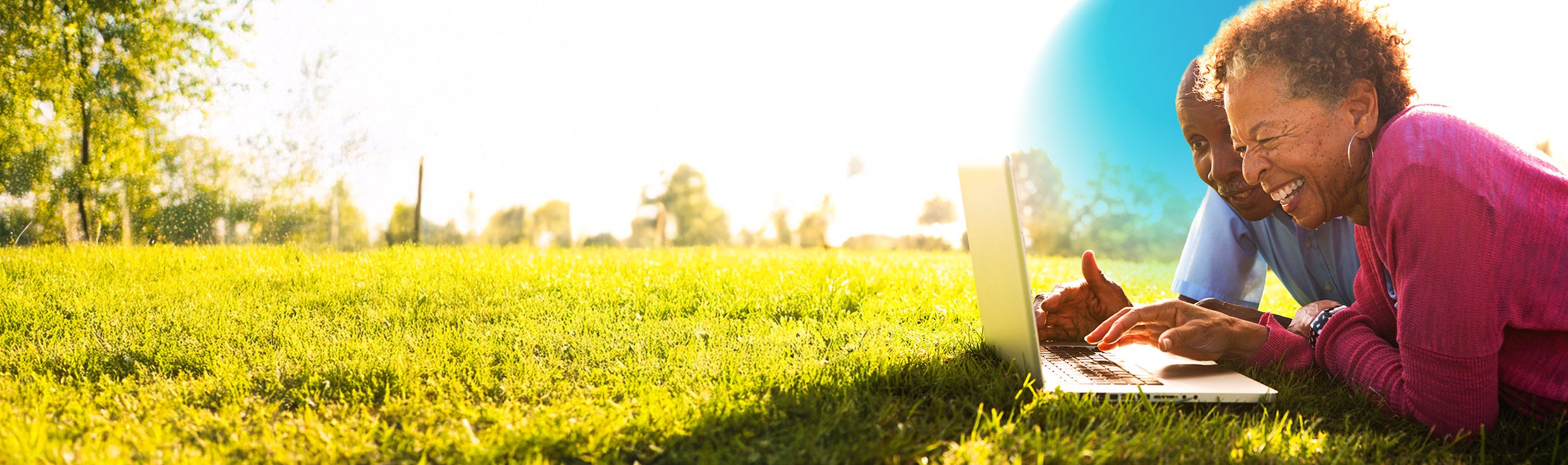 Senior African American woman wearing a red blouse with her husband in a  blue button up shirt lying on the grass smiling scrolling on their laptop 