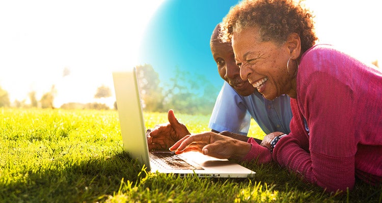 Senior African American woman wearing a red blouse with her husband in a  blue button up shirt lying on the grass smiling scrolling on their laptop 