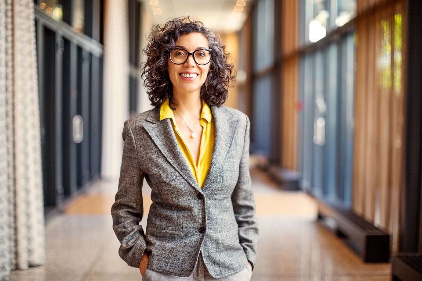 Middle aged female professor wearing a grey blazer and glasses with grey curly hair standing in a school hallway 