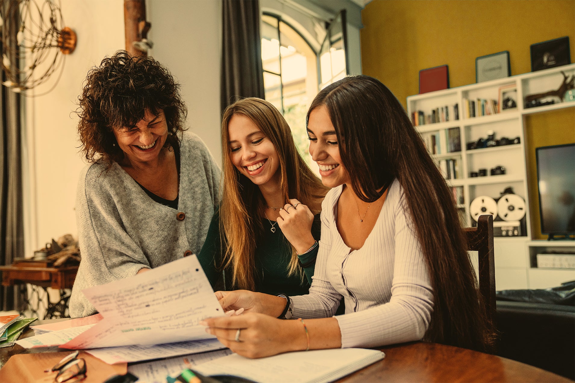 Two daughters smiling sitting at a kitchen table showing their mother a piece of paper 