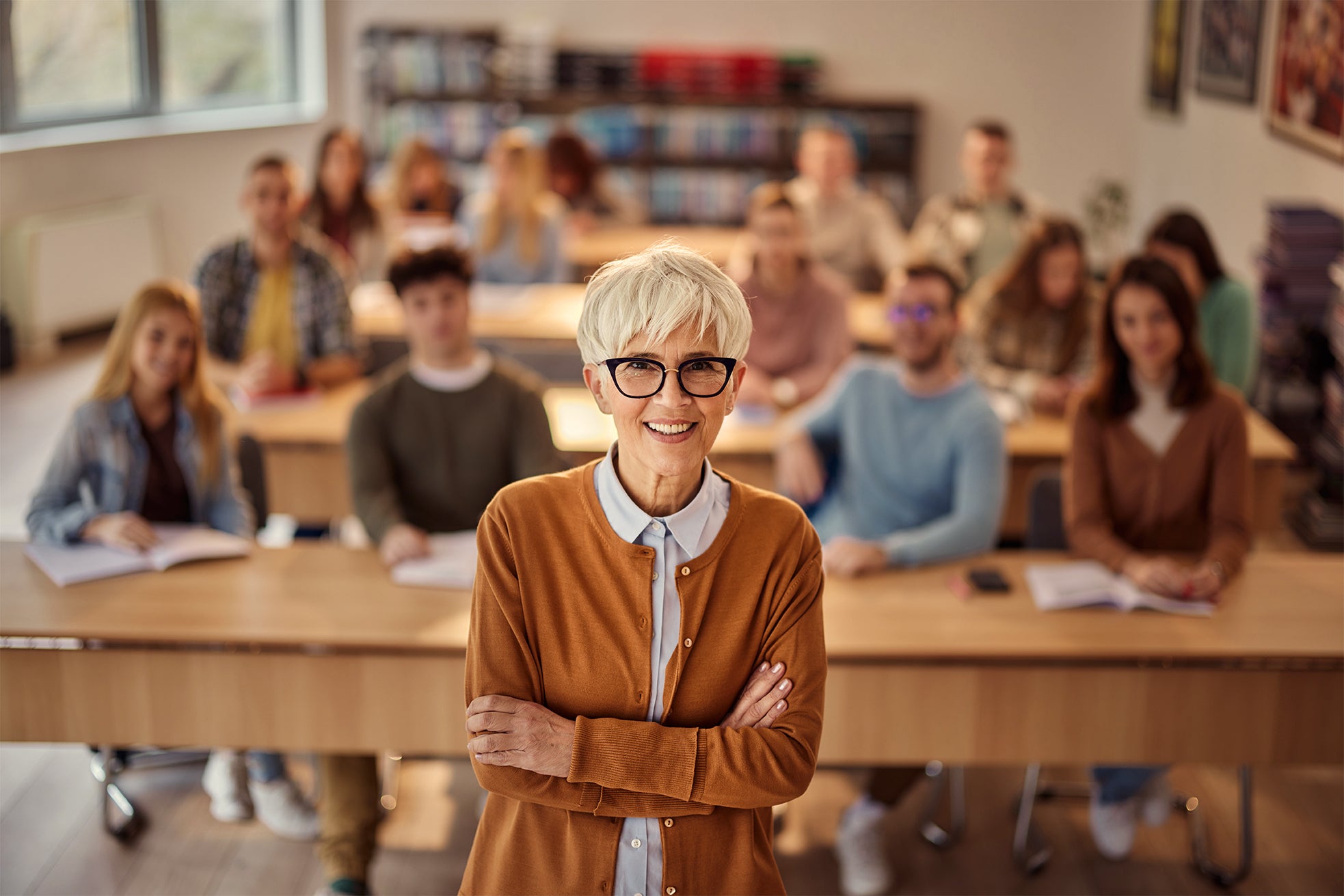 Senior Caucasian female professor wearing an orange cardigan standing in front of her classroom with students sitting at desks 