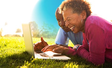 Older man and woman lying on the grass during a sunny day smiling and looking at laptop with blue circle hovering behind them