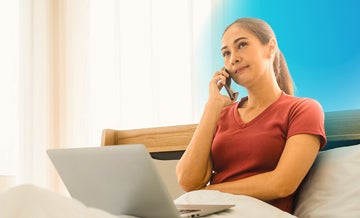 Woman sitting in bed talking on the phone with a laptop on her lap and blue circle hovering behind her 