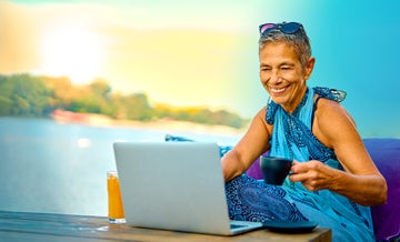 Older woman smiling and sitting by the water with a coffee mug in her hand and looking at her laptop