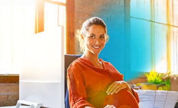 Smiling pregnant businesswoman sitting at computer desk with blue circled gradient hovering around her