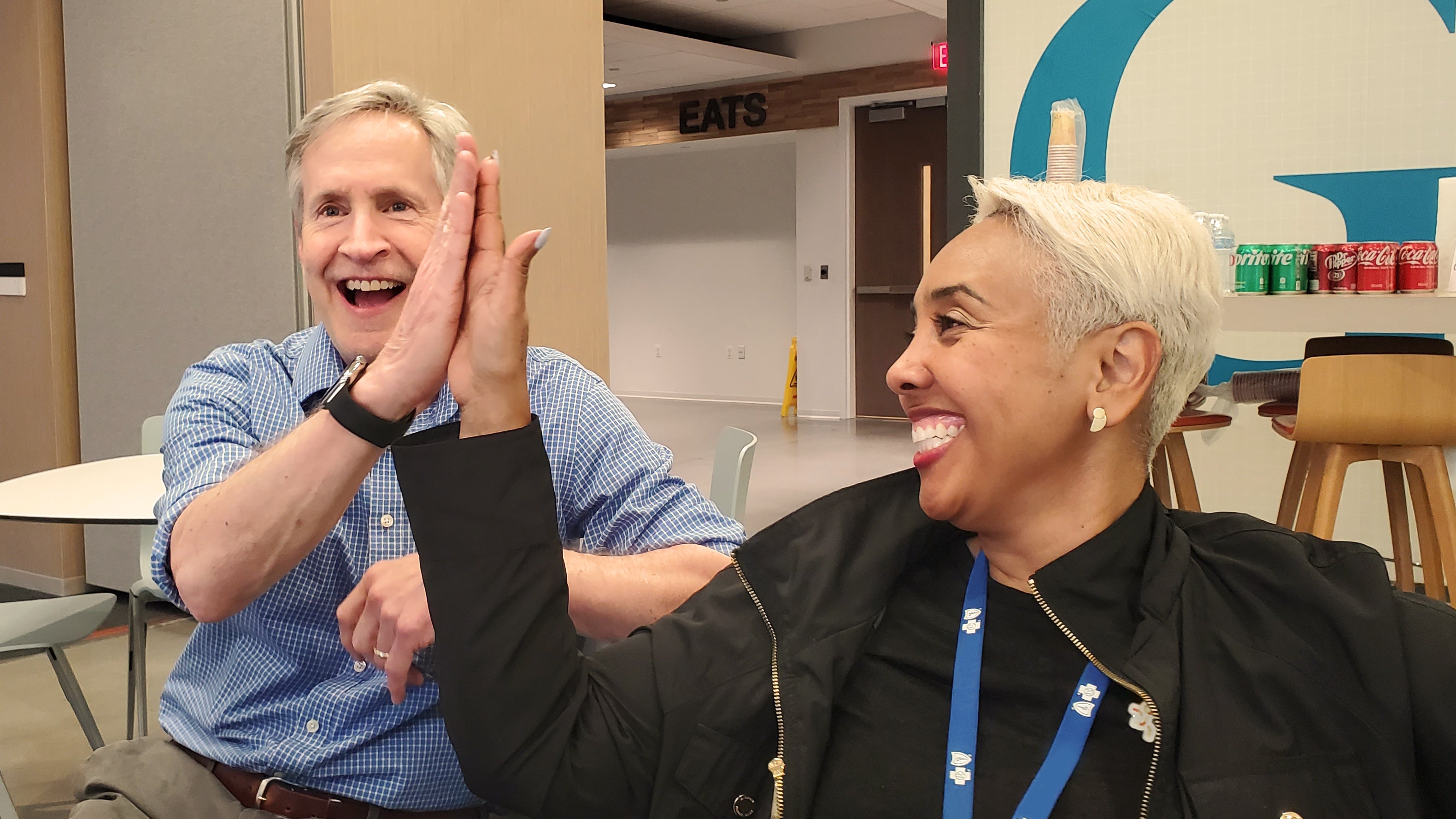 Two coworkers smiling and high fiving sitting in a cafeteria at an office 