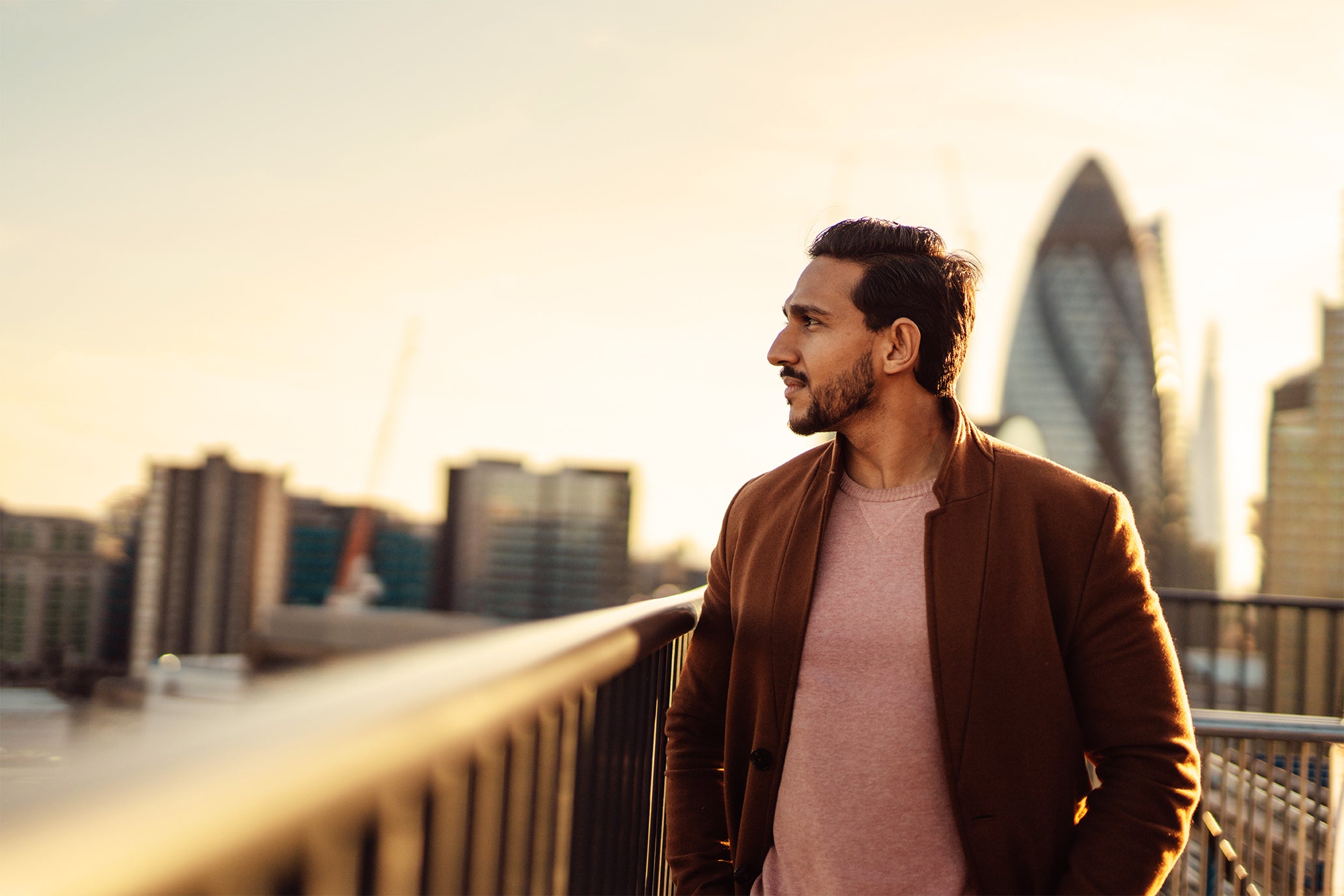 Man wearing a brown jacket overlooking the city skyline on a balcony 