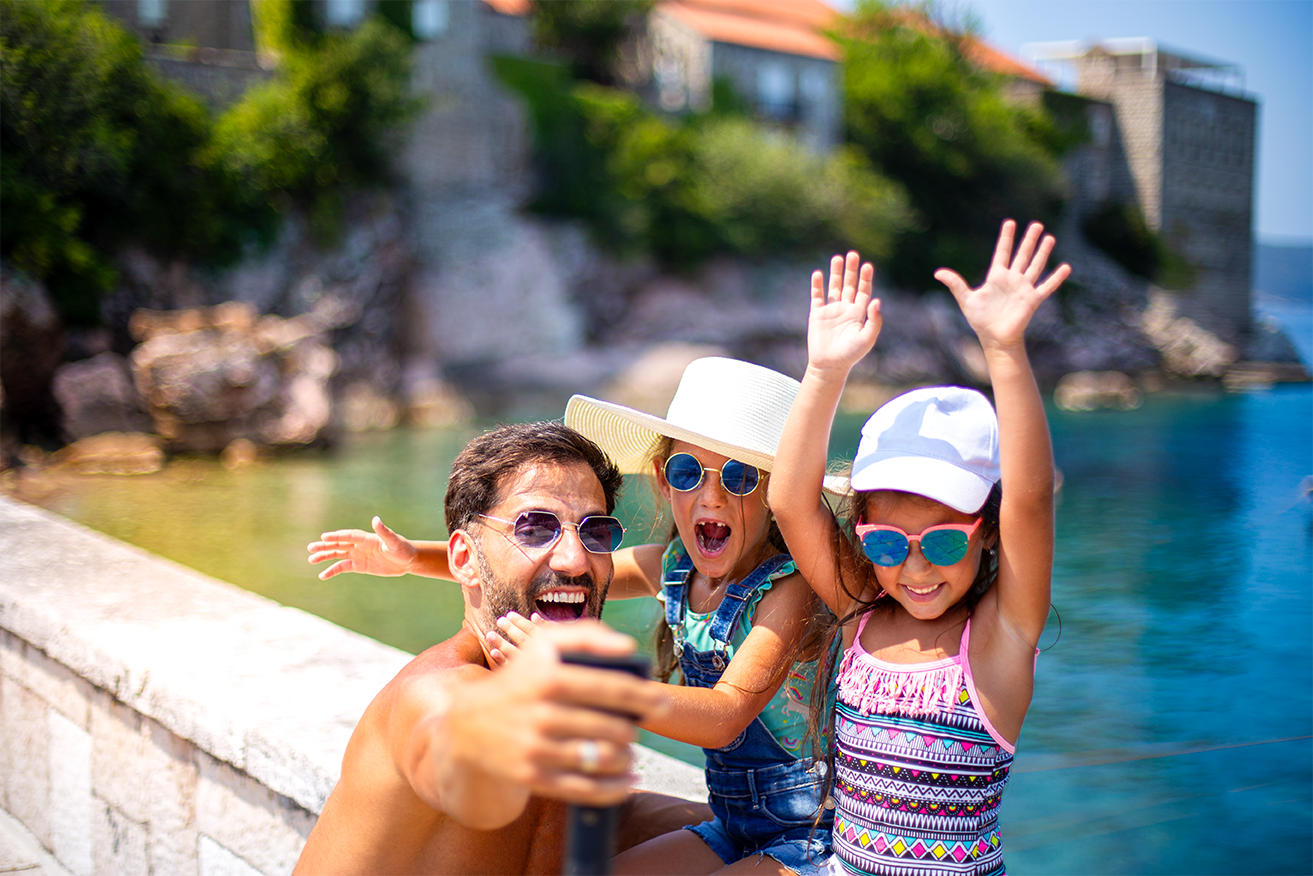 Father with two young child daughters all wearing sunglasses and swimwear taking a selfie and laughing by the water 