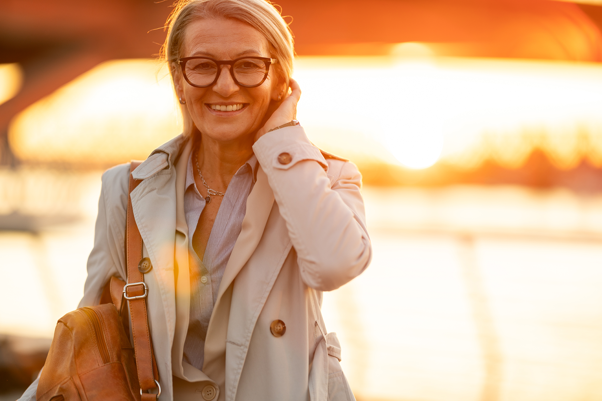 Smiling woman wearing a shoulder bag, glasses and jacket looking ahead with a sunset behind her