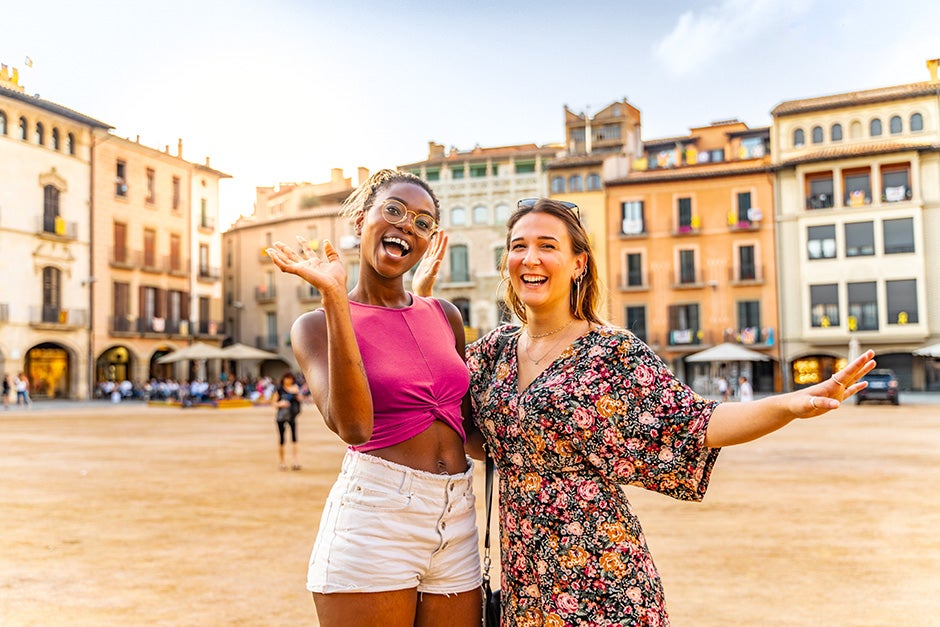 Two smiling women standing in front of shops in a European looking shopping and eating area 