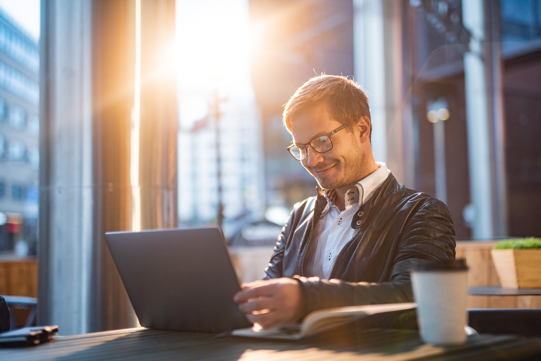 Caucasian man wearing glasses and a black leather jacket sitting at a desk by a window smiling and looking at his laptop 