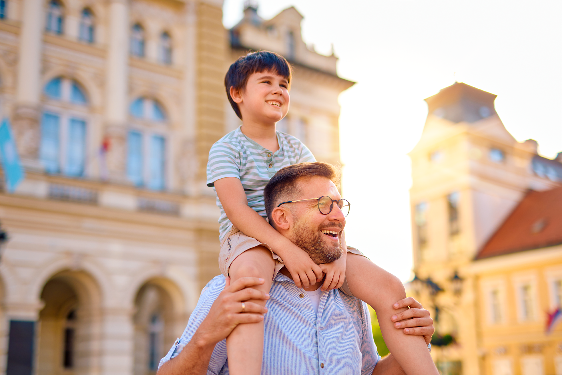 Caucasian father standing outside wearing glasses holding his young son on his shoulders who are both smiling 