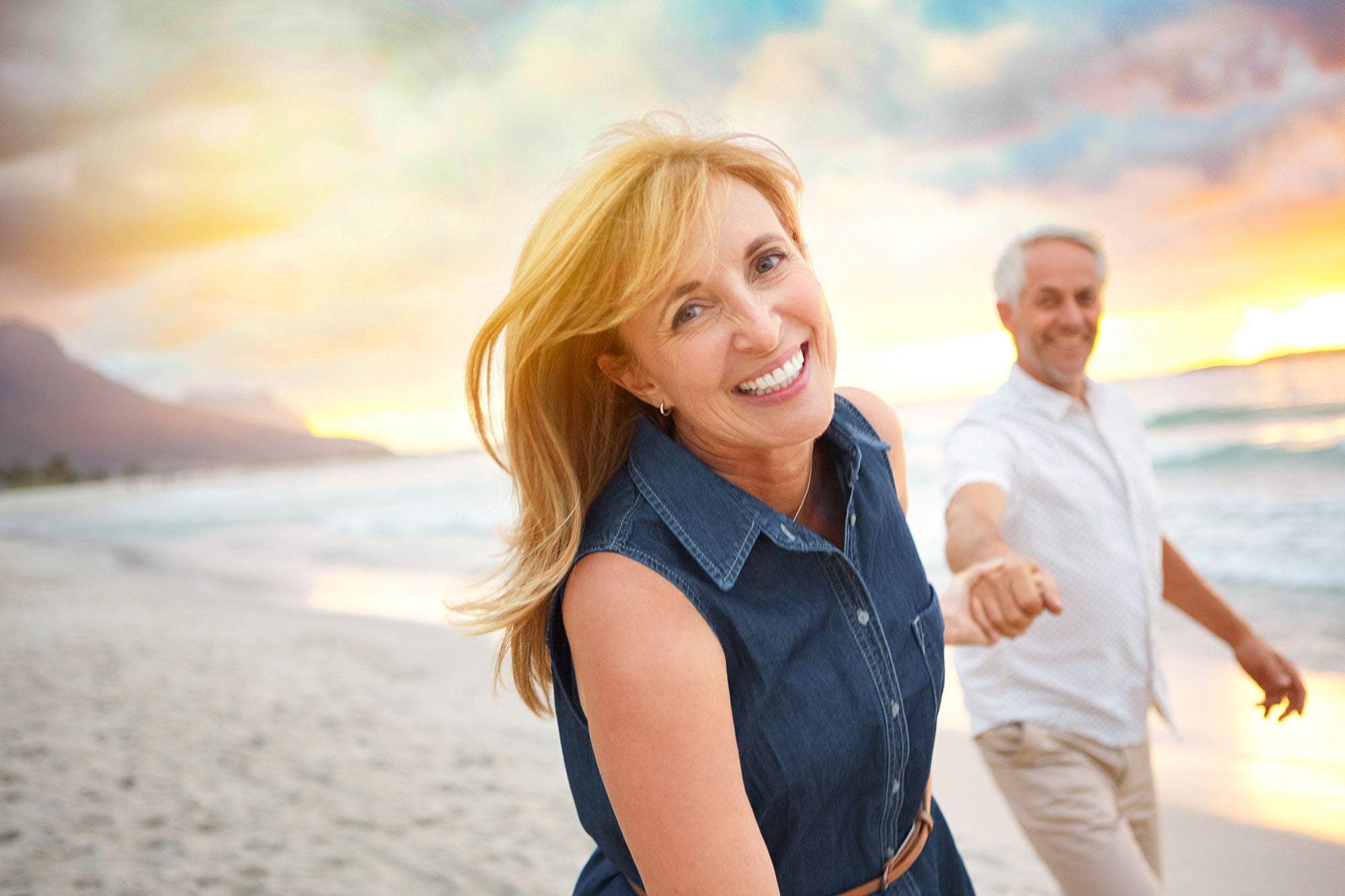 Middle aged white woman with blonde hair and a middle aged man holding hands smiling walking on a beach at sunset