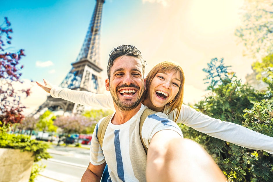 Young Caucasian man in a white and blue t-shirt and a Caucasian woman in a white shirt smiling in front of the Eifel Tower on a sunny day 