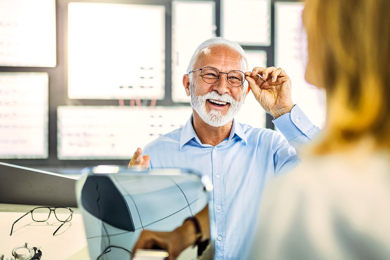 Senior man wearing glasses and a blue button up shirt smiling while seemingly trying on eyewear