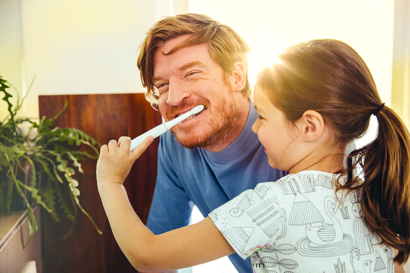 Caucasian smiling man wearing a blue shirt leaned over while young child daughter is brushing his teeth 